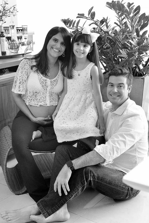 Imagem em pb da família, mãe a esquerda, filha sentada parcialmente no seu colo e o pai adireita delas, sentado ao chão'