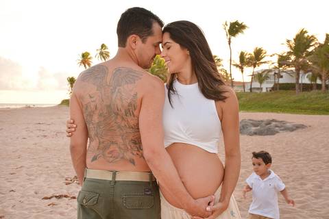 Na praia, o papai está de costas para câmera, mostrando uma enorme tatugame nas costas e abraçando a barriga da mãe, enquanto o irmãozinho corre ao fundo'