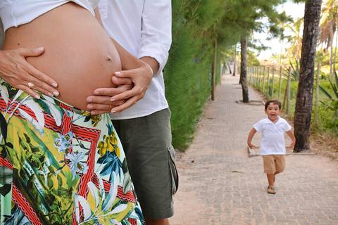 Papai abraça a barriga da mamãe enquanto o irmão corre ao fundo do jardim'