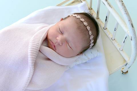 Foto em ângulo diferenciado, diagonal feita de cima da bebê enrolada numa manta rosa, dormindo numa mini cama'