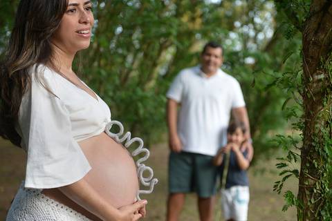 Foto da gestante com um arco bordado com a palavra AMOR sobre sua barriga e o filho e pai ao fundo'