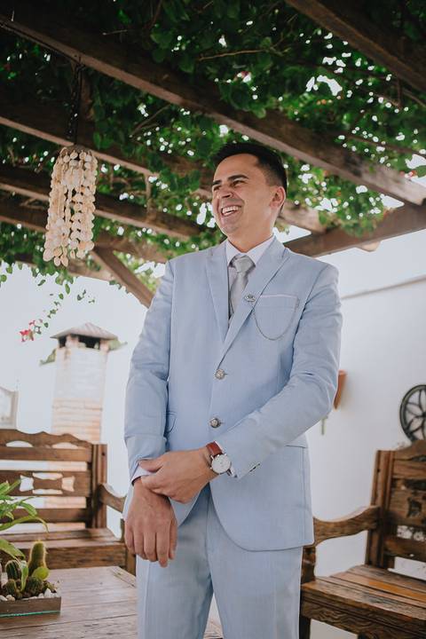 Noivo sorridente durante o making of, interagindo com outra pessoa enquanto ajusta o punho da camisa para o casamento.'