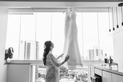 Foto em preto e branco da noiva admirando o vestido que se encontra pendurado na janela do apartamento'