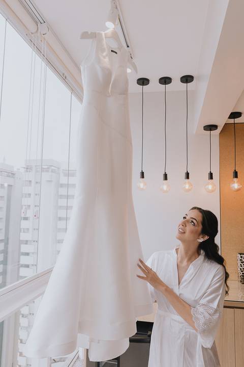 Foto da noiva admirando o vestido que se encontra pendurado na janela do apartamento'