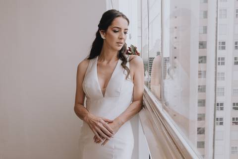 Foto da noiva noiva acabou de ficar pronta para o seu casamento e está posando para a foto, contemplando a vista de um prédio alto, no qual ela está,  usando um vestido muito bem desenhado, sóbrio, branco, sem pedrarias, perfeito para o estilo dela'