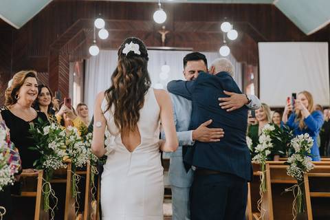 Foto da noiva entrando na capela Nossa Senhora Aparecida, com o pai da noiva abraçando o noivo'