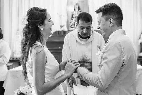 Foto do casal durante a cerimônia de matrimônio na Capela Nossa Senhora Aparecida, no momento da troca de alianças em preto e branco, com o casal sorrindo'