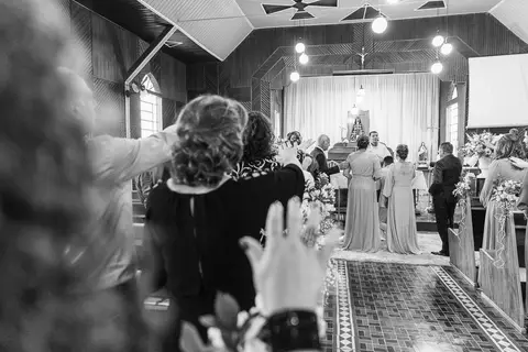 Foto do casal durante a cerimônia de matrimônio na Capela Nossa Senhora Aparecida, no momento da benção da família sobre o casal'