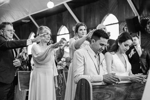 Foto do casal durante a cerimônia de matrimônio na Capela Nossa Senhora Aparecida, no momento da benção da família sobre o casal'