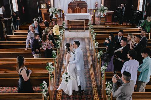 Foto do casal durante a cerimônia de matrimônio na Capela Nossa Senhora Aparecida, no momento em que eles estão saindo da igreja e se beijam no meio do corredor'