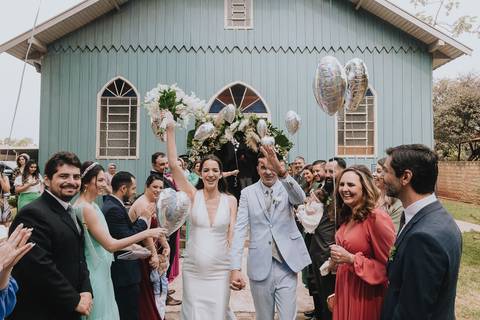 Foto do casal durante a cerimônia de matrimônio na Capela Nossa Senhora Aparecida, no momento em que eles estão saindo da igreja com os padrinhos comemorando'
