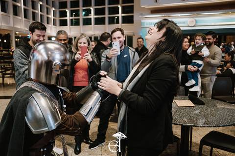 noiva surpreendida em pedido de casamento geek inspirado em star wars, the mandalorian, noivo ajoelhado segurando as alianças, vestido de mandaloriano'