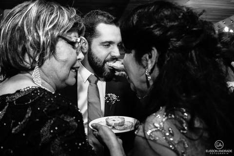 Casamento na chácara Carpe Diem, em são josé dos pinhais, curitiba, paraná, mãe do noivo servindo salgado na boca do noivo enquanto conversa com convidada'