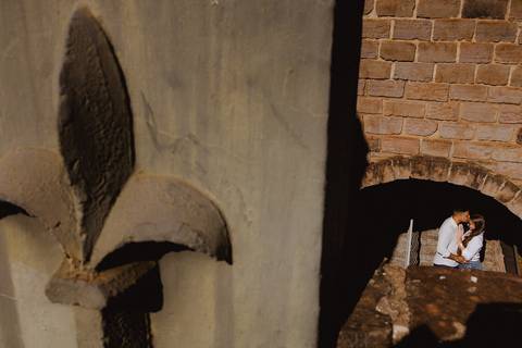 Casal em ensaio pré-wedding na Vinícola La Dorni em Bandeirantes, PR, visto através de elemento arquitetônico em pedra, criando composição artística e clima intimista'