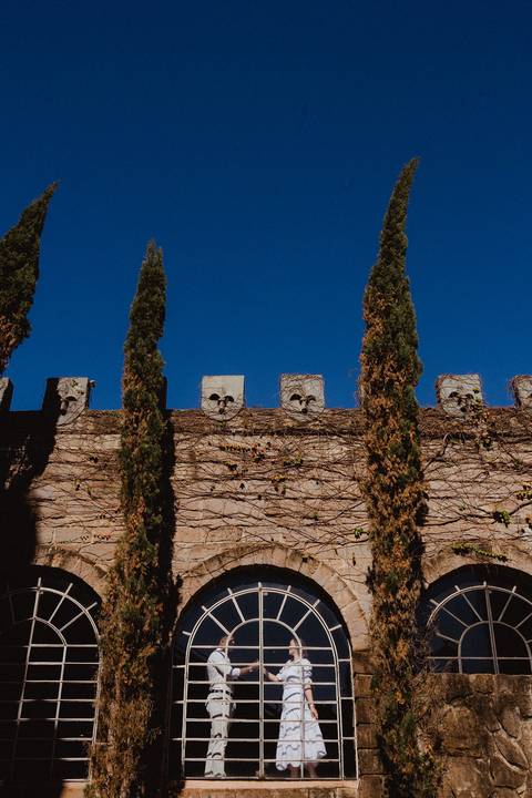 Casal em ensaio pré-wedding na Vinícola La Dorni em Bandeirantes, PR, posando dentro dos arcos de pedra cobertos por trepadeiras, sob céu azul intenso e composição vertical artística'