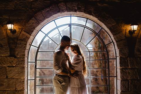 Casal em ensaio pré-wedding na Vinícola La Dorni em Bandeirantes, PR, abraçado diante da janela arqueada de pedra iluminada pela luz natural, em cena romântica e acolhedora'