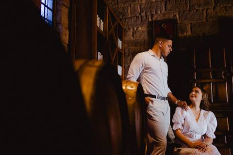 Casal em ensaio pré-wedding na Vinícola La Dorni em Bandeirantes, PR, em ambiente interno rústico com tonéis e paredes de pedra, trocando olhares em clima romântico'