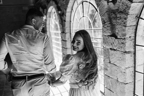 Casal em ensaio pré-wedding na Vinícola La Dorni em Bandeirantes, PR, caminhando de mãos dadas entre janelas arqueadas de pedra, com a noiva sorrindo para a câmera em foto em preto e branco'