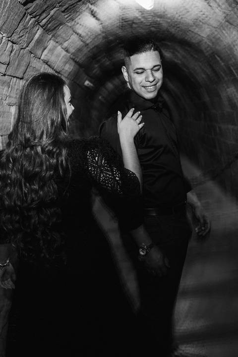 Casal em ensaio pré-wedding na Vinícola La Dorni, no túnel de pedras, com ela tocando o ombro dele enquanto ele sorri; fotografia em preto e branco com blur art intencional e luz dramática para clima intimista'