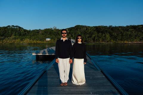 Arthur e Mari em terceiro figurino preto e branco, retrato simétrico no píer ao entardecer, óculos escuros'