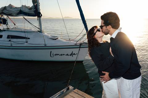 Arthur e Mari se beijando ao entardecer ao lado do veleiro Limonada, luz dourada refletindo na água'
