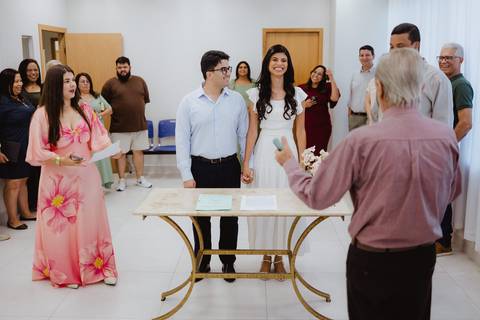 Noivos ao lado de uma testemunha sorrindo para o celebrante'