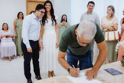 Testemunha assinando o documento enquanto os noivos observam sorrindo'