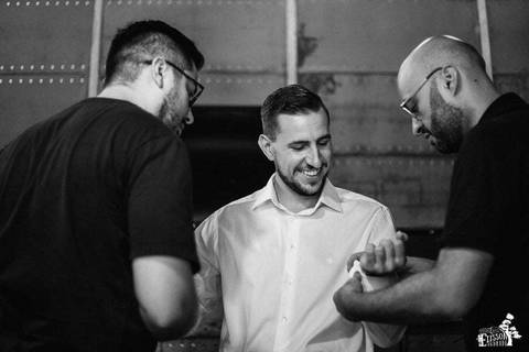 padrinhos ajudando o noivo a dobrar e abotoar a camisa branca, na barbearia, enquanto o noivo se arruma para o casamento'