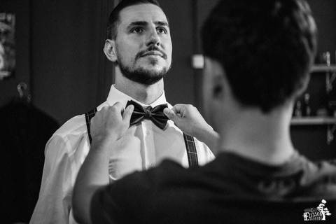 padrinho arrumando a gravata borboleta do noivo, na barbearia, enquanto o noivo se arruma para o casamento'