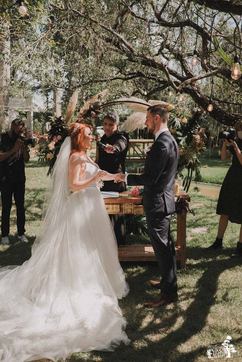 Troca de alianças durante cerimonia de casamento ao ar livre, com decoração boho, em frente a um arco de flores e galhos verdes'