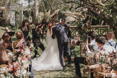 beijo dos recém casados durante cerimonia de casamento ao ar livre, com decoração boho, em frente a um arco de flores e galhos verdes'