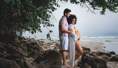 gestante nas pedras na praia e marido abraçando'