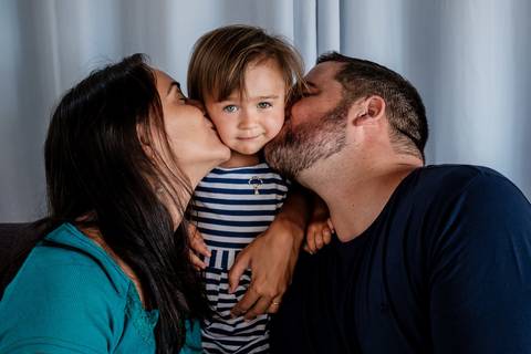 pai e mãe beijando a filha na bochecha e a menina olha para a câmera'