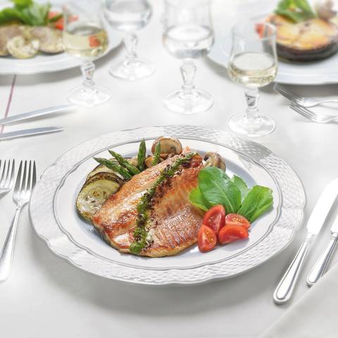 Fotografía publicitaria de José Chiodini de un plato elegante de pescado acompañado de espárragos, tomate y berenjenas grilladas, servidos en mesa gourmet con copas de vino.'