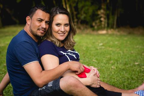 gestante sorridente com o seu marido sentados em um gramado e ele segura um par de sapatinho vermelho sobre a barriga dela'