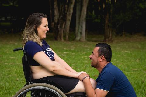 gestante na cadeira de roda de mão dadas com seu marido ajoelhado em sua frente e os dois sorrindo'