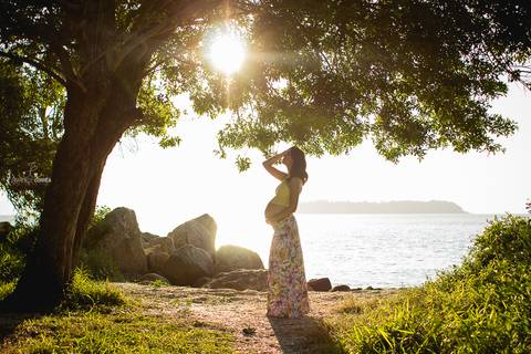 Foto da gestante perto de uma árvore entrando uma luz linda do sol'