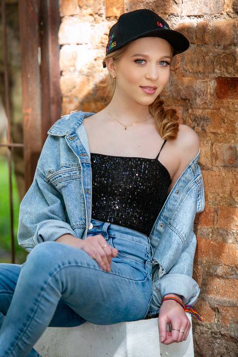 book de 15 anos menina loira com roupas jeans e blusa preta de paetê sentada na calçada com parede rustica ao fundo usado boné. Fábio Martins fotografo '