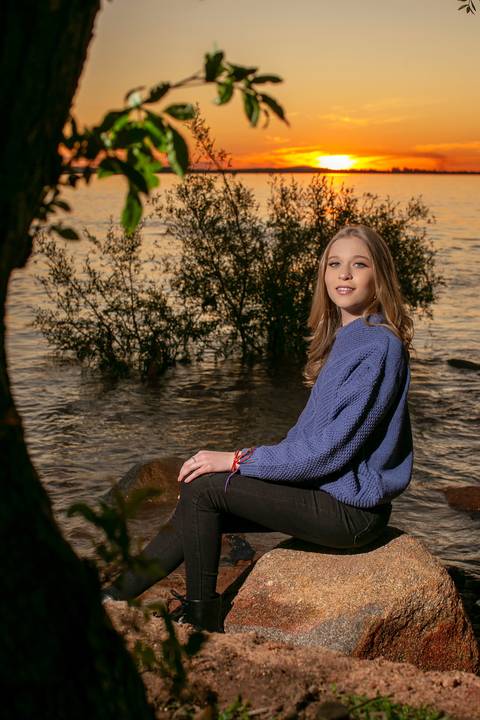 menina calça preta blusa azul com tênis vans ao por do sol na orla do Guaíba sentada em um pedra. Fábio Martins fotografo '