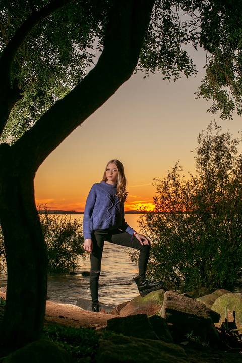 menina calça preta blusa azul com tênis vans ao por do sol na orla do Guaíba com árvore do lado esquerdo. Fábio Martins fotografo  '