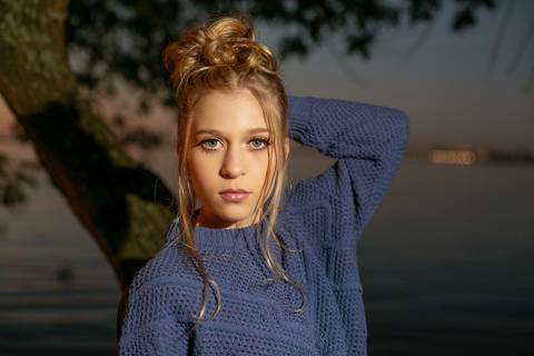 menina calça preta blusa azul com tênis vans ao por do sol na orla do Guaíba  encostada em um árvore. Fábio Martins fotografo'