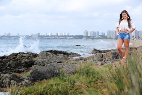 Fotografia-de-Street-Book-de-15-anos-na-praia-de-Punta-del-Este-em-Maldonado-no-Uruguai-modelo-na-praia-de-punta-del-este-foto-na-praia-de-punta-del-este-fundo-praia-com-predios-modelo-posando-para-foto-de-blusa-branca-e-jeans-pedras-beira-praia-punta-del'