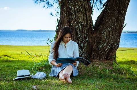 Ensaio fotográfico ao ar livre no campo, no Yacht Club em Araçatuba, SP. Ela esta sentada no gramado sob uma árvore ao fundo tem o rio Tietê. Ela lê um livro. '