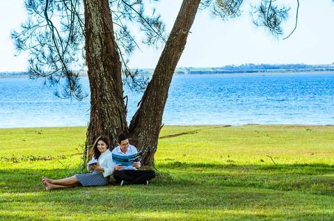 Ensaio fotográfico ao ar livre no campo, no Yacht Club em Araçatuba, SP. O casal esta sentada no gramado sob uma árvore ao fundo tem o rio Tietê. Eles lêem um livro. '