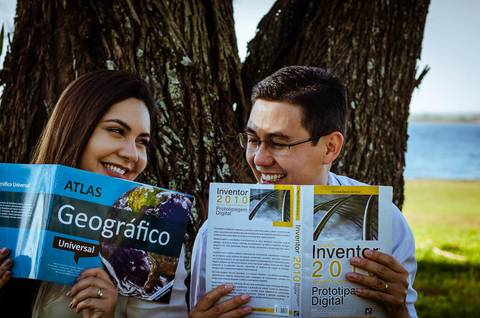 Ensaio fotográfico ao ar livre no campo, no Yacht Club em Araçatuba, SP. Eles estão se olhando e sorrindo enquanto seguram um livro cada um. Estão sentados sob uma árvore e ao fundo tem o rio Tietê. '