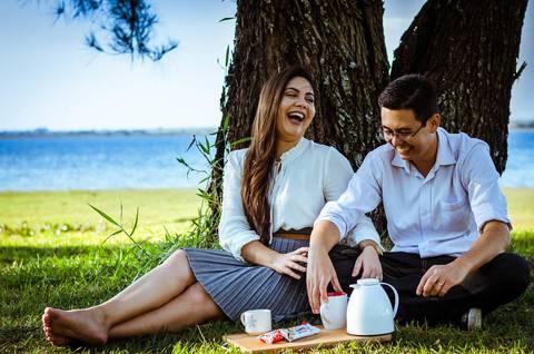 Ensaio fotográfico com um casal dando risadas. Estão sentados sob uma árvore frondosa em um gramado, ao fundo tem o rio Tietê. Eles estão comendo chocolate e tomando café.'