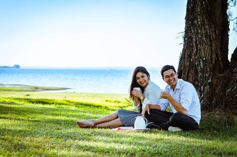 Ensaio fotográfico com um casal dando risadas. Estão sentados sob uma árvore frondosa em um gramado, ao fundo tem o rio Tietê. Eles estão comendo chocolate e tomando café.'