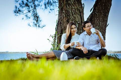 Ensaio fotográfico com um casal. Estão sentados sob uma árvore frondosa em um gramado, ao fundo tem o rio Tietê. Eles estão comendo chocolate e tomando café.'