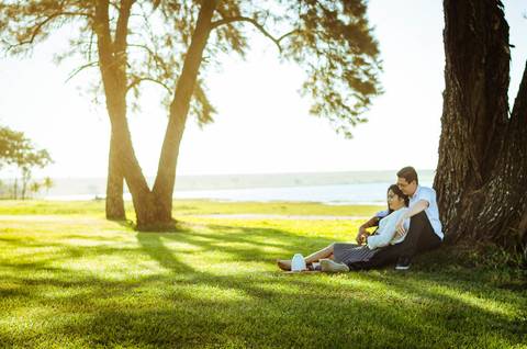 Ensaio fotográfico com um casal dando risadas. Estão sentados sob uma árvore frondosa em um gramado, ao fundo tem o rio Tietê. e várias outras árvores'
