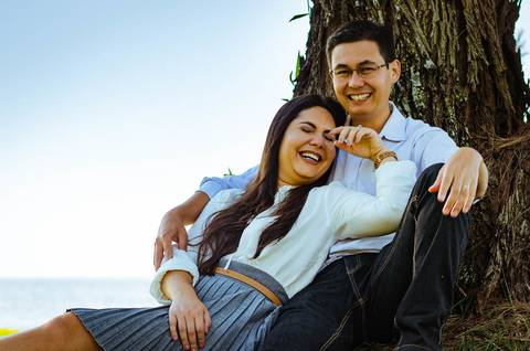Ensaio fotográfico do casal. Esta foto eles se abraçam com carinho sentindo uma brisa suave. O casal está dando risada, se divertindo.'